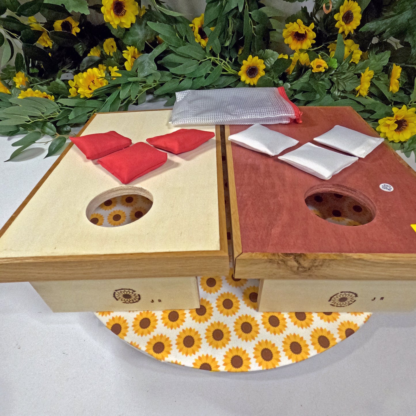 Wooden cornhole set with red and white boards on a white surface with sunflower pattern, surrounded by sunflowers.